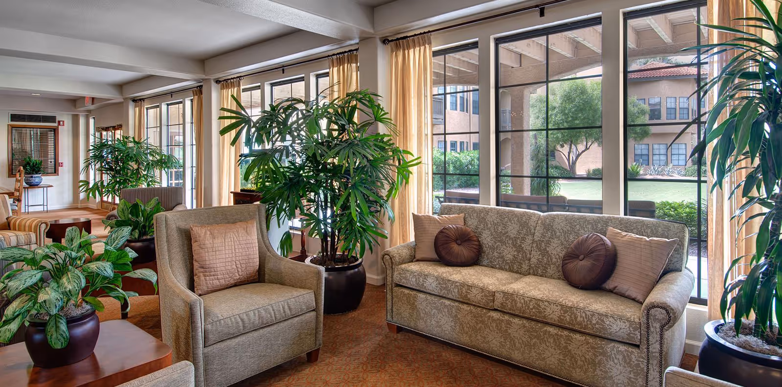 A cozy living room area with a patterned sofa and armchair, both adorned with decorative pillows. Large windows with beige curtains let in natural light and offer a view of a green outdoor courtyard. Several potted plants are placed around the room, adding a touch of greenery to the space.