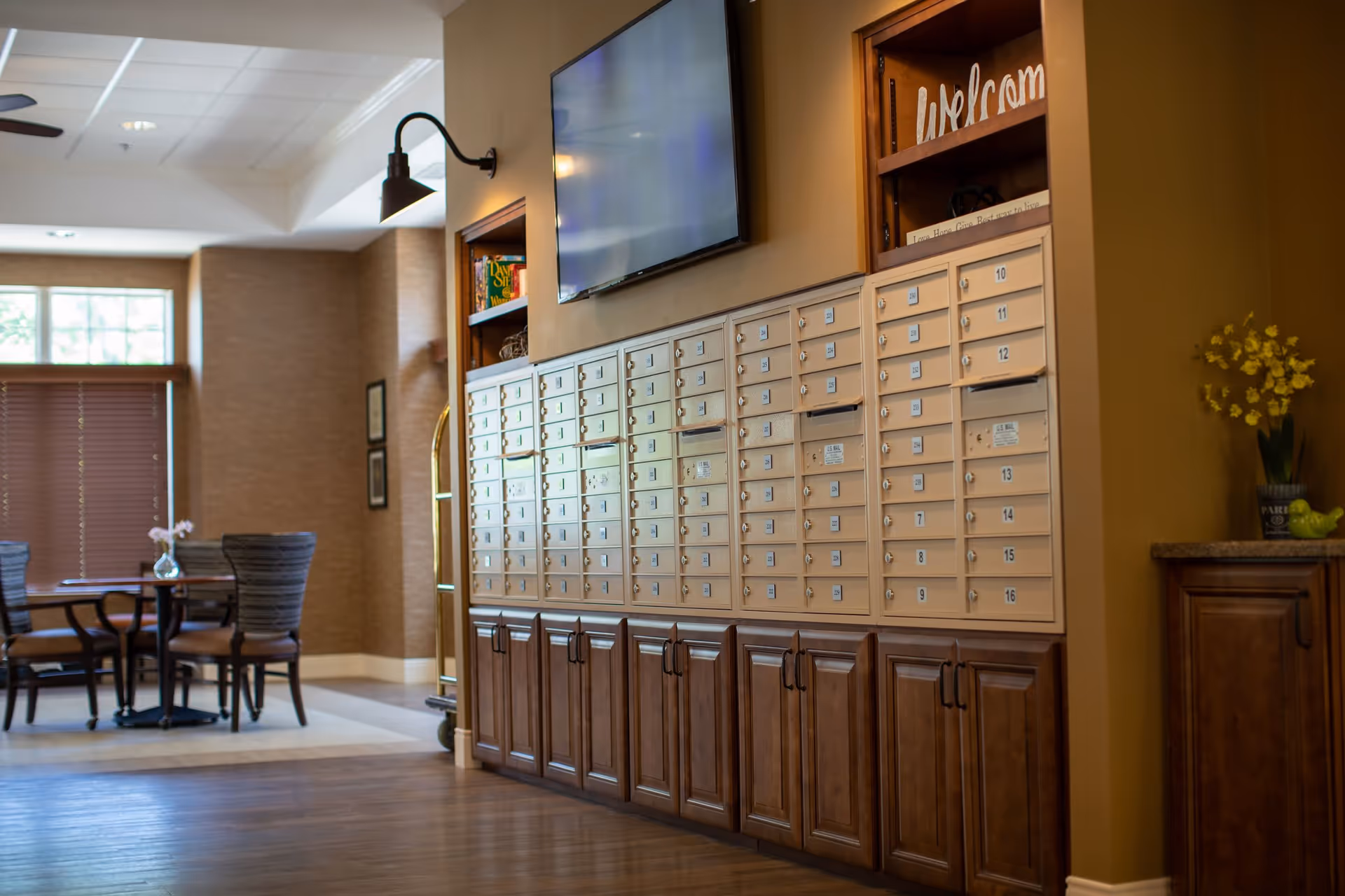 Interior view of a senior living facility lobby area featuring a wall of mailboxes with numbered slots and wooden cabinets below. Above the mailboxes is a mounted flat-screen TV. To the left, there is a seating area with a round table and chairs near a window with blinds. The space is warmly lit and decorated with a small vase of flowers and a welcome sign on a shelf.
