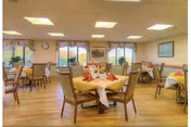 A well-lit dining room with multiple round tables covered with yellow tablecloths and set with red napkins. The room has large windows with floral curtains, wooden flooring, and framed artwork on the walls.