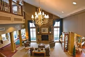 Spacious and elegant living room area with high vaulted ceilings, a large stone fireplace centered between two tall windows, a chandelier hanging from the ceiling, and comfortable seating including sofas and chairs arranged around a coffee table. The room features warm lighting, decorative curtains, and a wooden railing on the upper level overlooking the space.