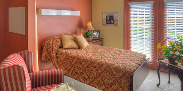 A cozy nursing-home bedroom with a patterned bed, striped armchair, nightstand with a lamp and photos, and French doors with blinds letting in light.