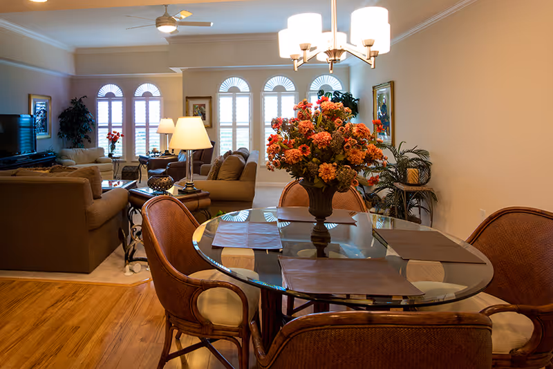 A cozy living and dining area with a round glass dining table set with four chairs and a large floral centerpiece. The living room features multiple cushioned sofas, a TV, lamps, and large arched windows with white shutters letting in natural light. The room has wooden flooring near the dining area and carpet in the living space, with neutral-colored walls and ceiling fans.