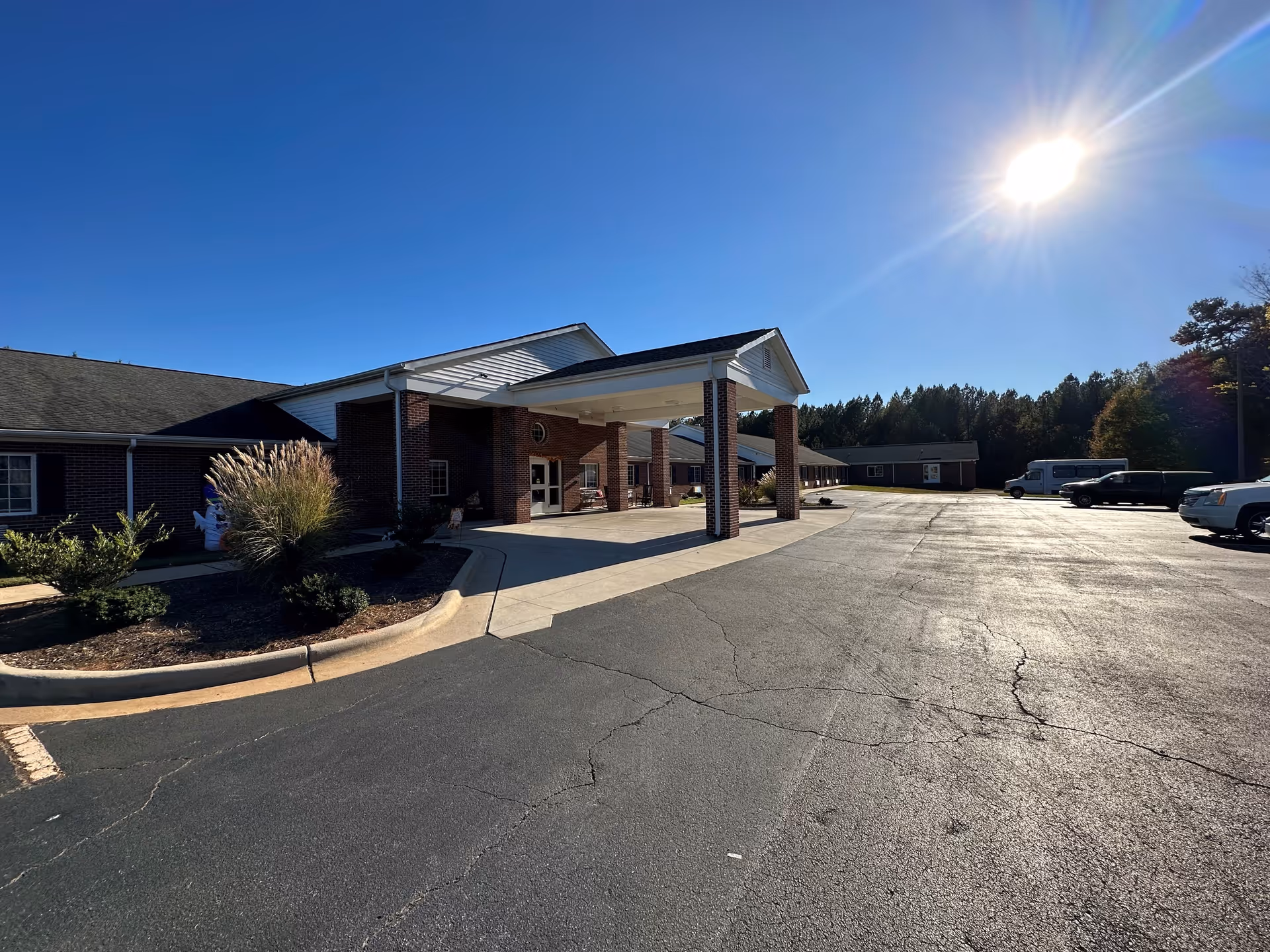 Exterior view of Wexford House facility showing a brick building with a covered entrance, a large paved parking area, some parked vehicles, and clear blue sky with the sun shining brightly.