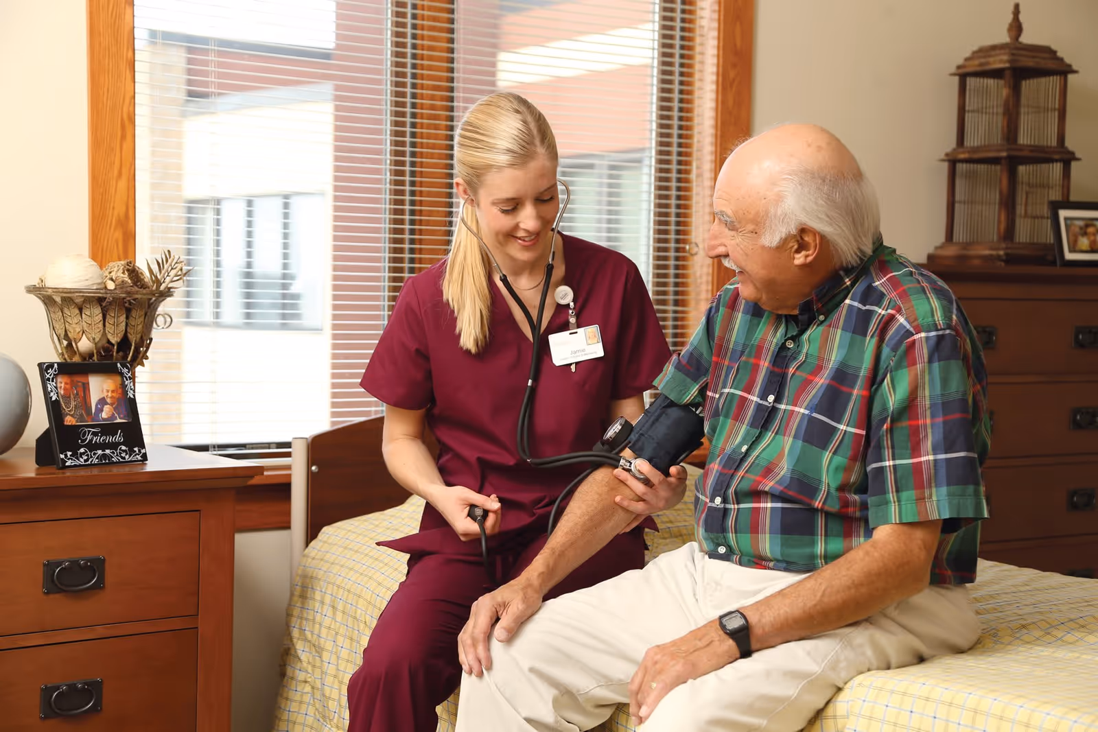 A female healthcare worker in burgundy scrubs is smiling while taking the blood pressure of an elderly man sitting on a bed in a cozy room with wooden furniture and a window with blinds.