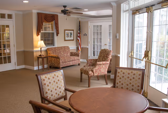 A bright communal seating area with upholstered chairs, a sofa, a round wooden table, a lamp, and glass doors to the outside.
