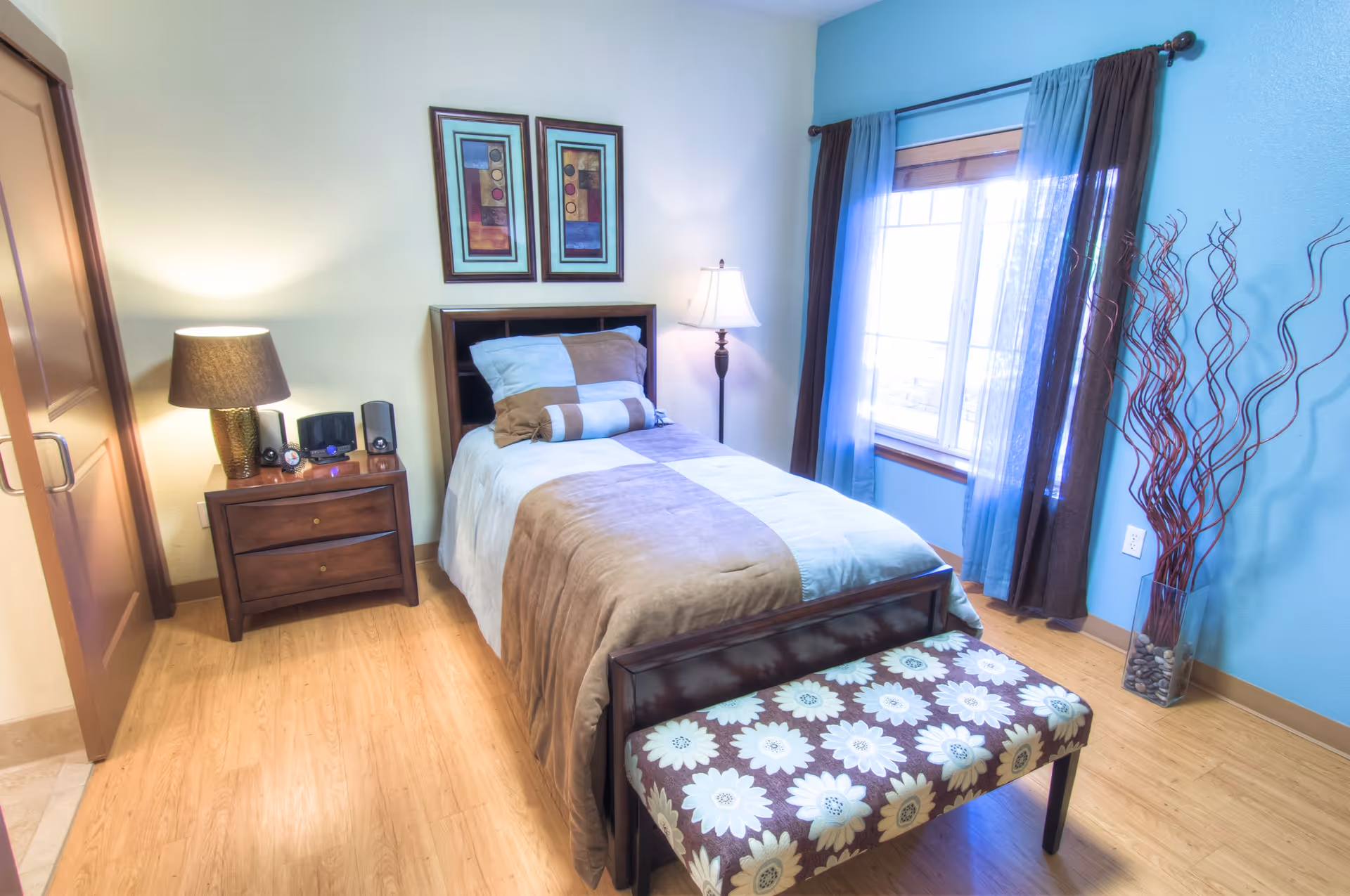 Neatly furnished single bedroom with a bed, nightstand, lamp and a window with blue curtains.