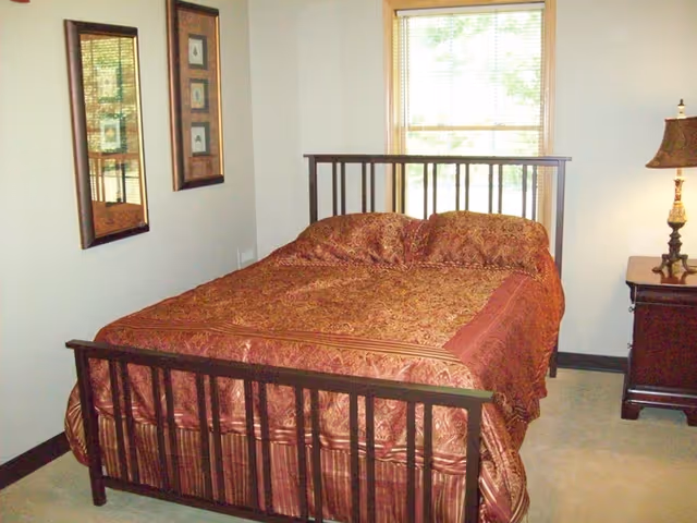 A small bedroom with a wooden bed frame and patterned red bedding, a nightstand with a lamp, two framed pictures on the wall, and a window.