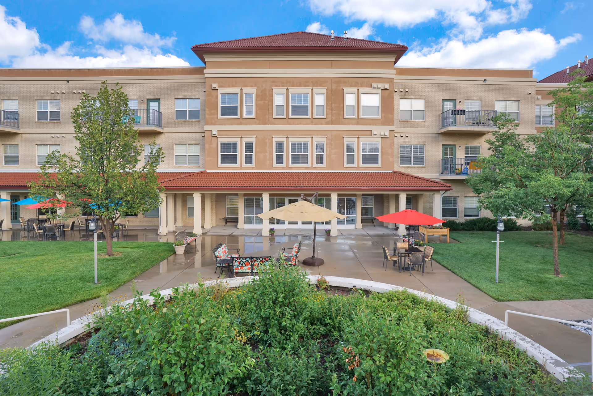 Exterior view of a multi-story senior living facility with a red-tiled roof and beige walls. The foreground features a landscaped garden with green shrubs and trees, a concrete patio with tables, chairs, and umbrellas in red, beige, and blue colors. The sky is blue with some clouds.