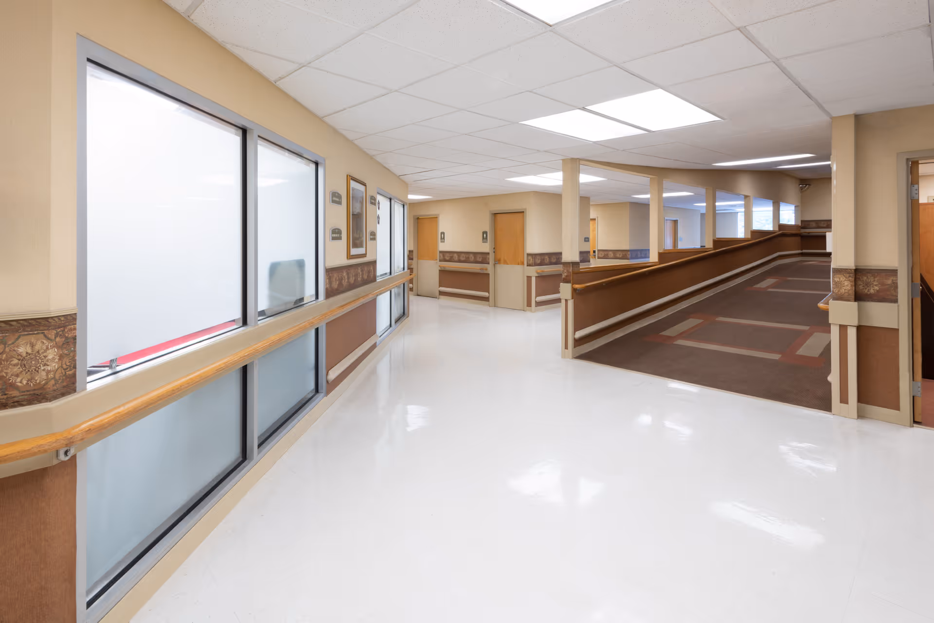 Interior hallway of Langhorne Gardens Health & Rehabilitation Center with white shiny floors, beige walls, handrails, and a carpeted ramp leading to another section. The hallway has several doors and windows with frosted glass panels.