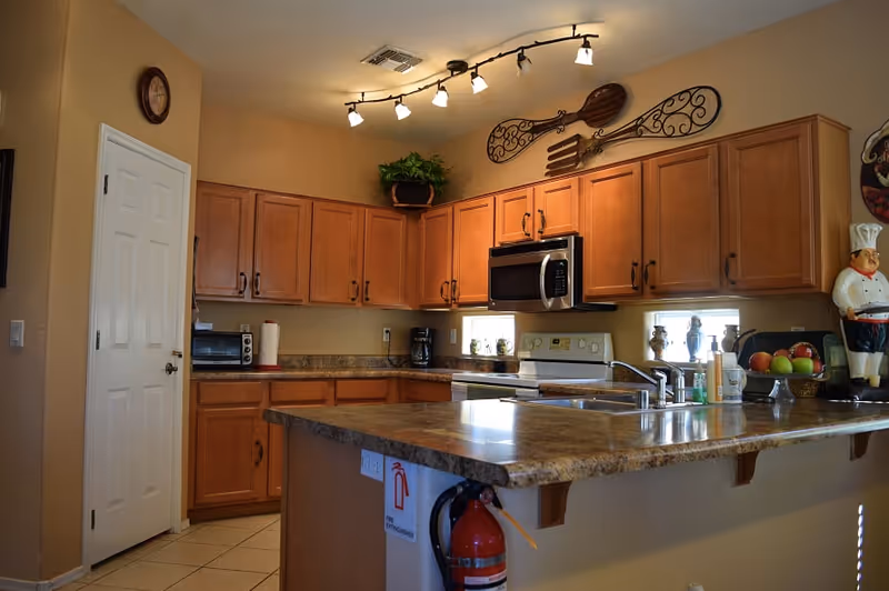 A kitchen with wooden cabinets, a countertop with a sink, a microwave, a stove, a coffee maker, and various kitchen decorations including a chef figurine and a bowl of fruit. There is a fire extinguisher mounted on the side of the counter and decorative wall art above the cabinets.