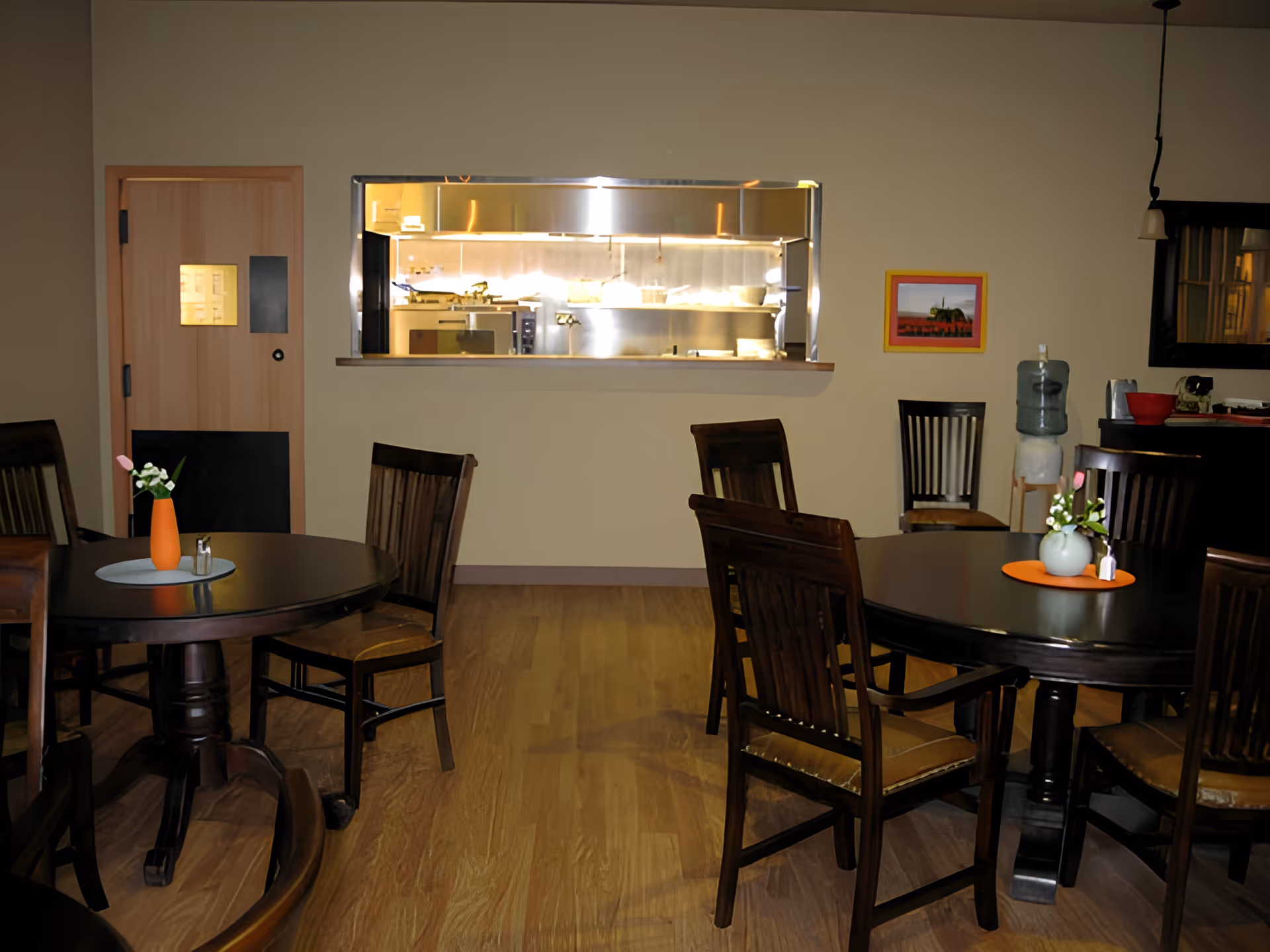 Interior view of a dining area with round wooden tables and chairs. Each table has a small vase with flowers. There is a serving window in the wall showing a stainless steel kitchen area behind it. A wooden door is on the left side of the wall, and a water dispenser and framed picture are on the right side.