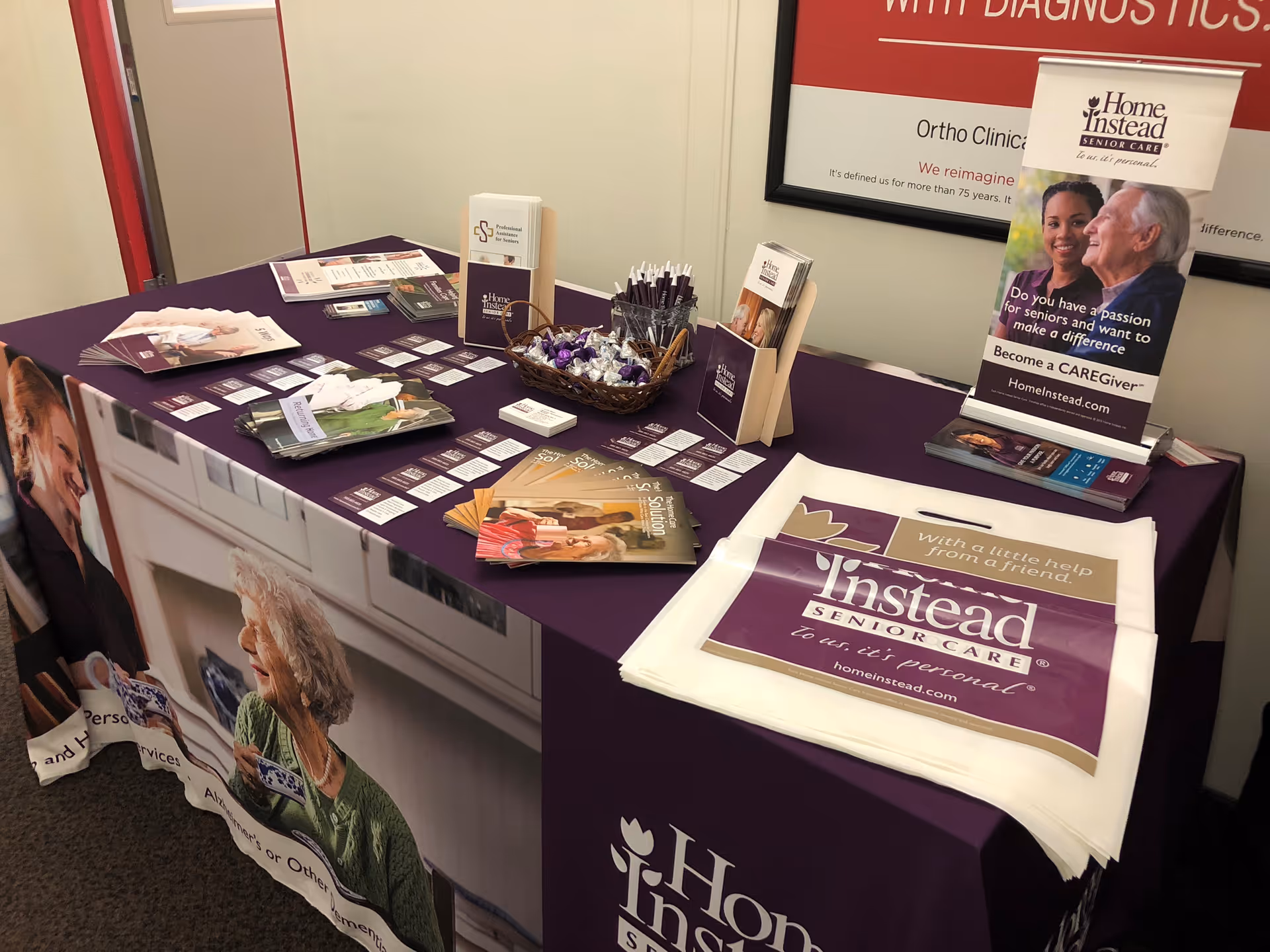 A purple-clothed table inside an office displaying Home Instead Senior Care brochures, pens, candy, and branded tote bags.