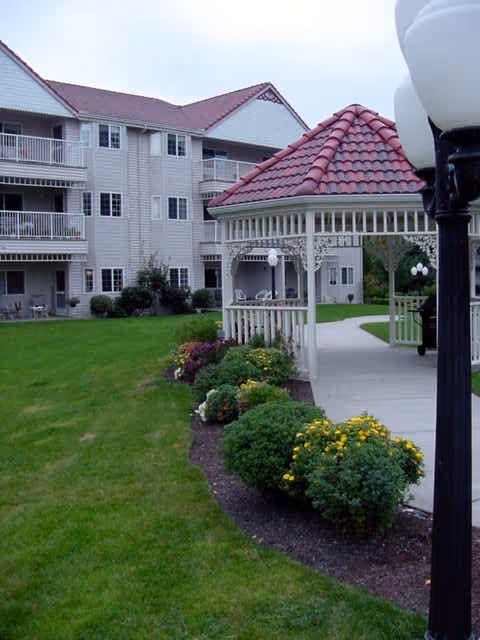 Outdoor view of Riverview Terrace showing a well-maintained lawn with flower beds, a white gazebo with a red tiled roof, and a multi-story residential building with balconies in the background.
