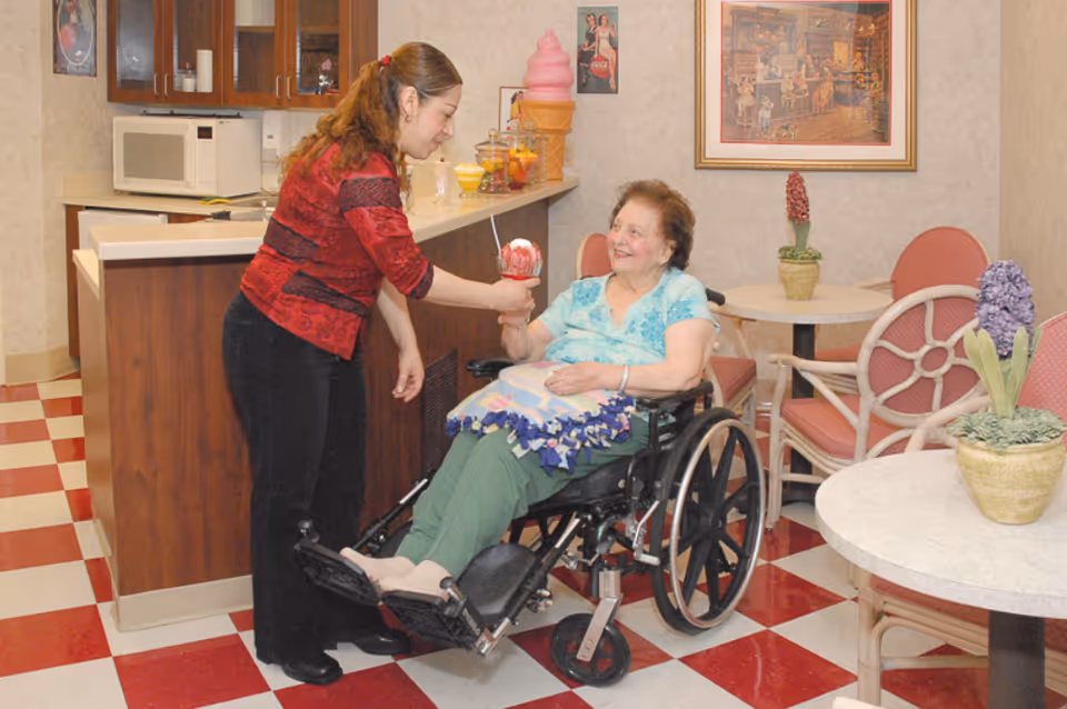 A woman in a red and black top is handing a dessert cup with a spoon to an elderly woman sitting in a wheelchair in a room with red and white checkered floor tiles. The room has a counter with a microwave and various items, round tables with pink chairs, and potted plants. A framed picture hangs on the wall.