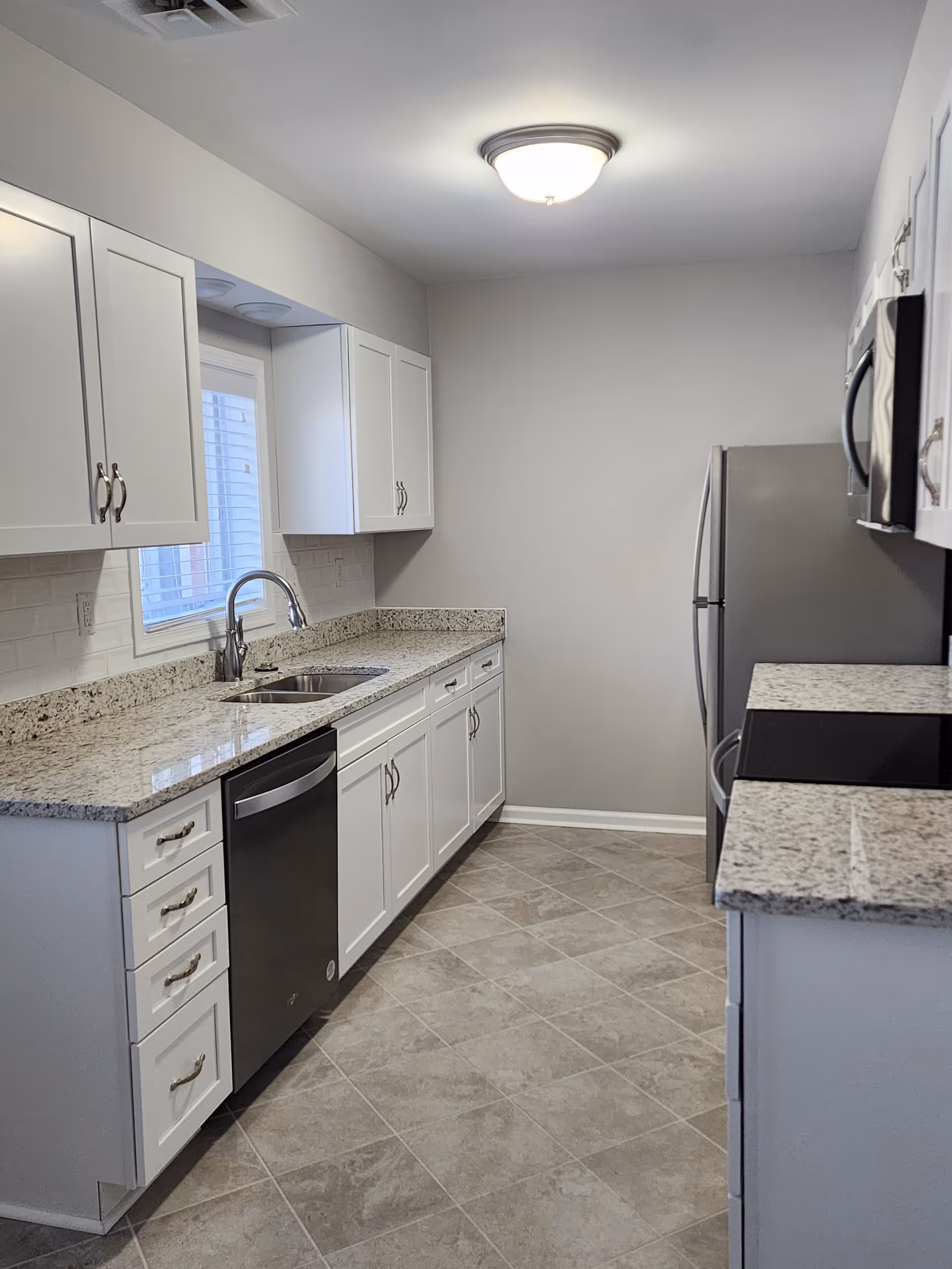A modern kitchen with white cabinets, granite countertops, a stainless steel dishwasher, refrigerator, and microwave. The floor is tiled with a light gray pattern, and there is a window above the sink allowing natural light in.