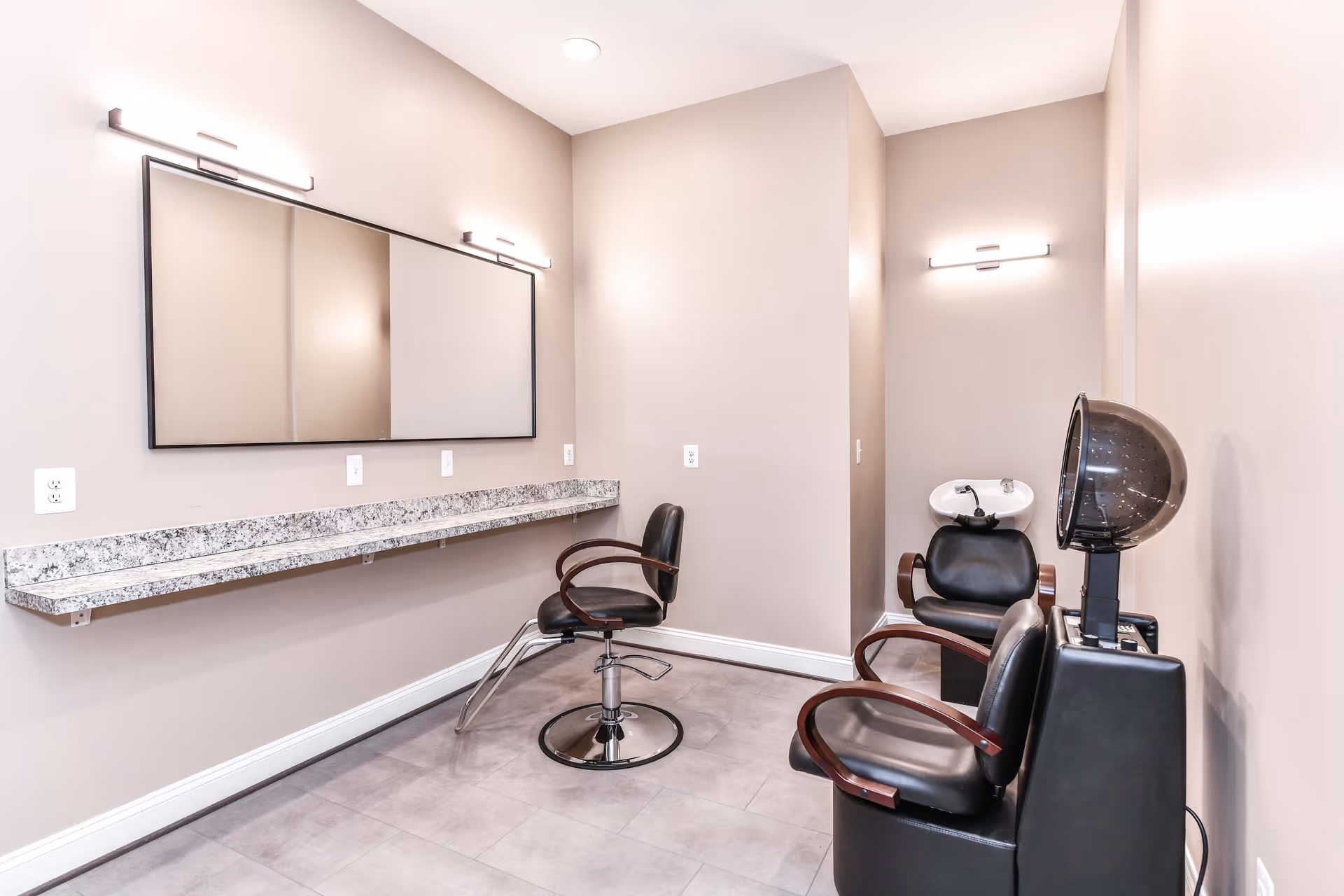 A small salon room with two black salon chairs, one with a hair dryer hood attached and the other positioned in front of a long granite countertop with a large mirror above it. The walls are painted beige and the floor is tiled in gray. There is a hair washing station with a black chair and white sink in the corner.