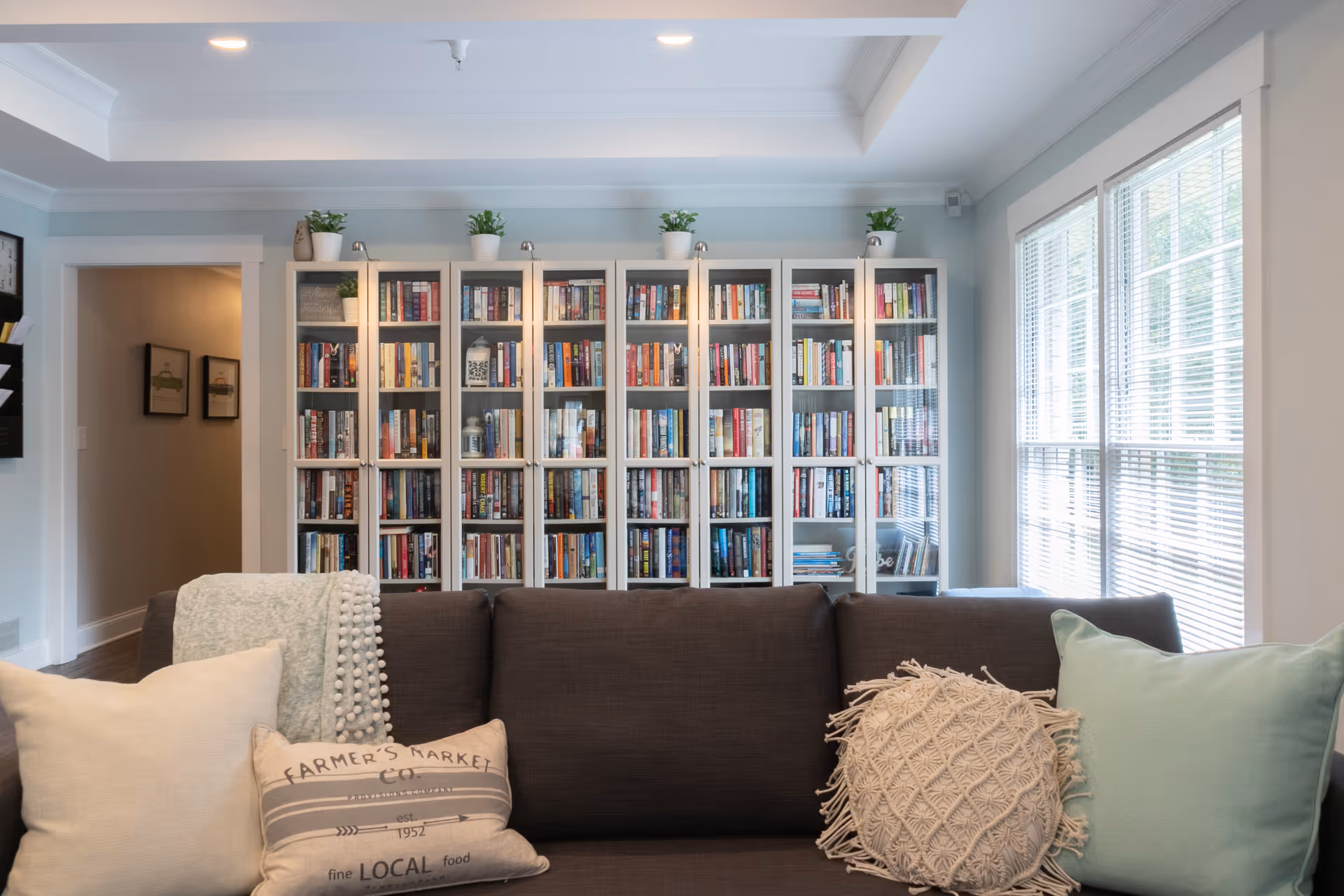 A cozy living room with a dark gray sofa adorned with various decorative pillows and a throw blanket. Behind the sofa is a large white bookshelf filled with numerous books and some small potted plants on top. To the right, large windows with blinds allow natural light to fill the room. The walls are painted light blue, and there is a doorway leading to another room on the left.
