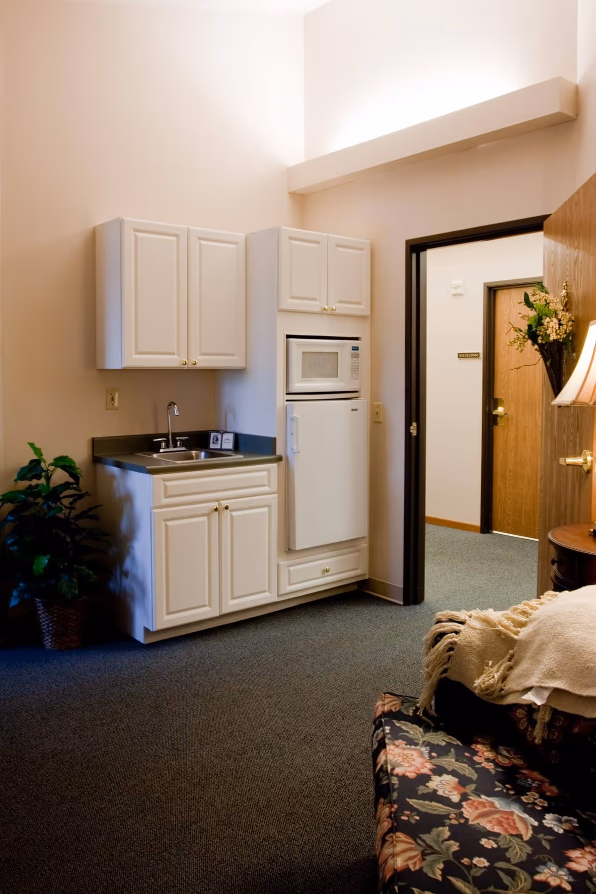 Small kitchenette with white cabinets, sink, microwave and refrigerator in a carpeted senior living suite, with a floral sofa and open doorway.