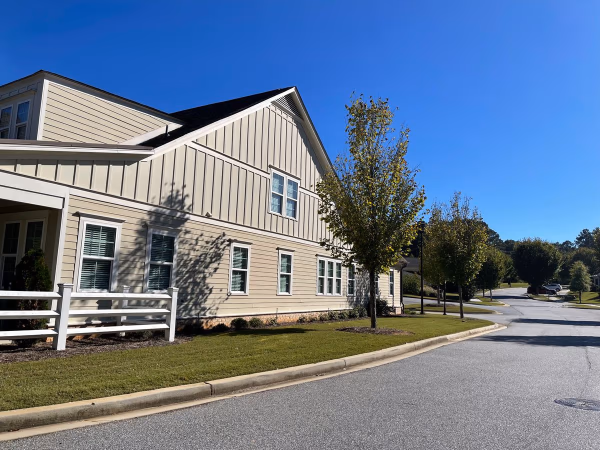 Sunlit beige-sided retirement community building with windows, a white fence, young trees and a curved road under a clear blue sky.