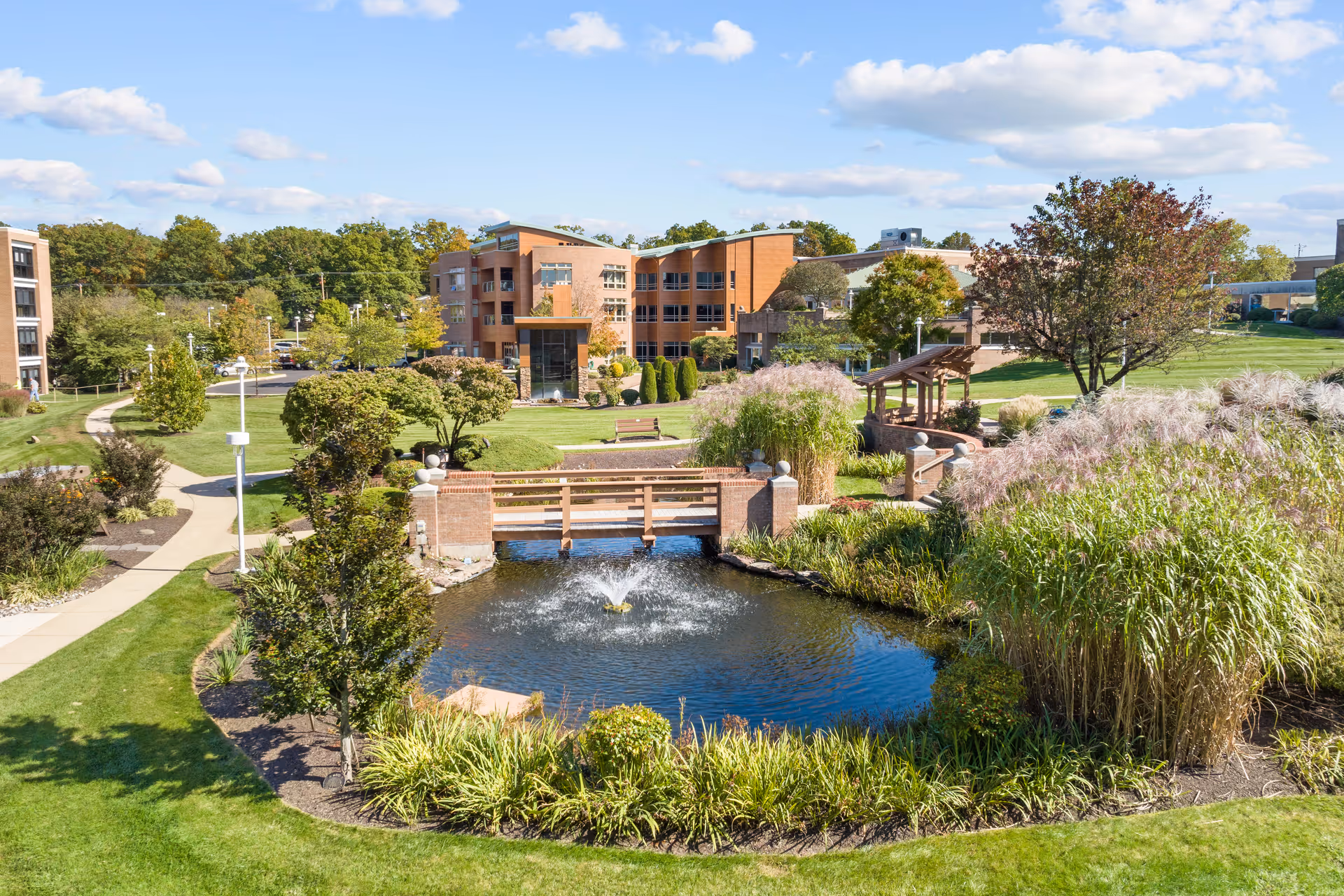 A landscaped outdoor area at The Community at Rockhill featuring a small pond with a fountain, a wooden bridge, walking paths, benches, trees, and ornamental grasses under a partly cloudy sky.