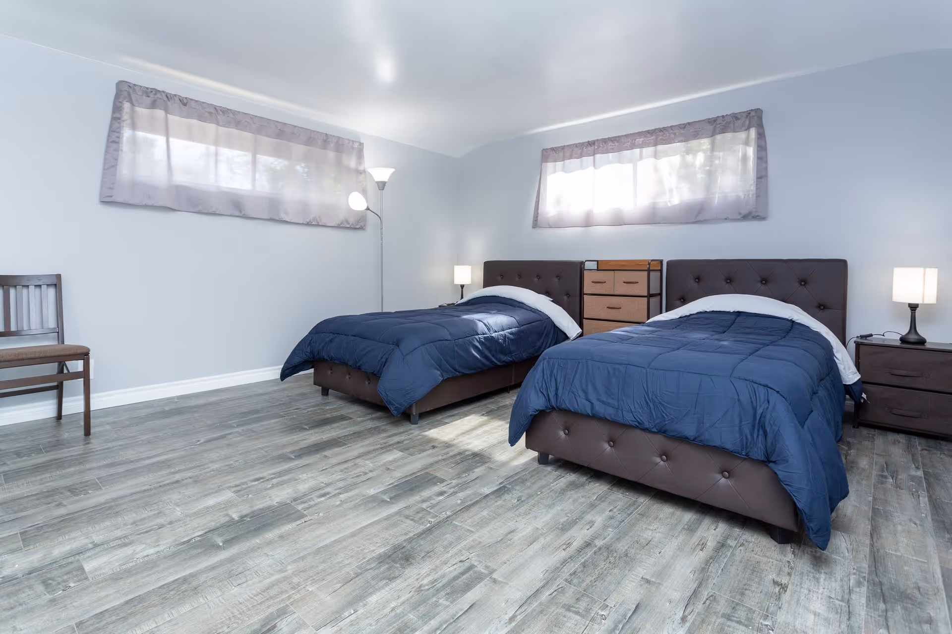 A bright bedroom with two twin beds featuring dark brown tufted headboards and navy blue comforters. Between the beds is a small wooden dresser with drawers, and on either side of the beds are nightstands with lamps. The room has light gray walls, two windows with sheer curtains, a wooden chair in the corner, and a floor lamp providing additional light. The floor is covered with light-colored wood laminate.