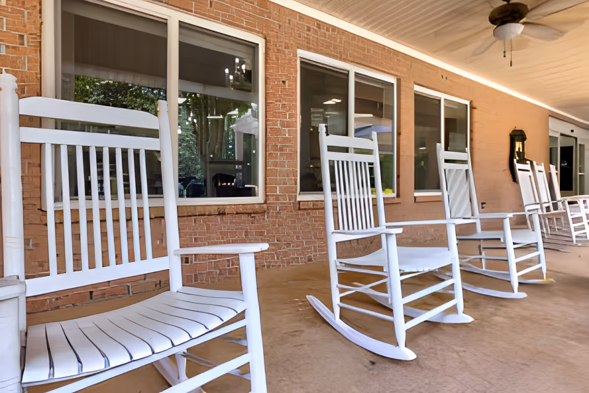 A row of white wooden rocking chairs lined up on a covered porch with a brick wall and several windows in the background. A ceiling fan is mounted above, and the porch floor is a smooth concrete surface.