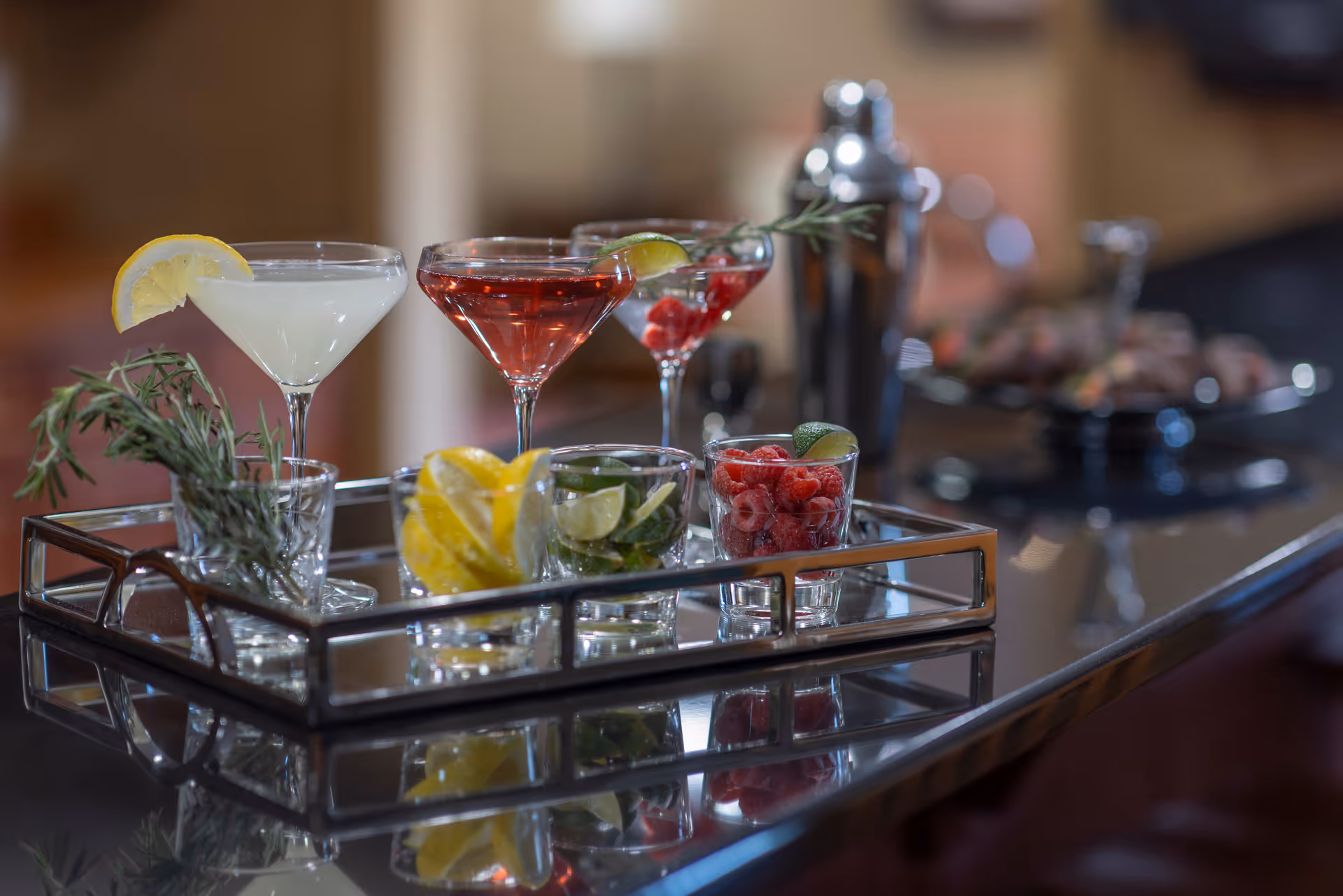 A tray on a reflective countertop holding two cocktail glasses with lemon and lime garnishes, a glass with raspberries, and small glasses containing lemon slices, lime slices, and rosemary sprigs. A cocktail shaker and a blurred plate of appetizers are visible in the background.