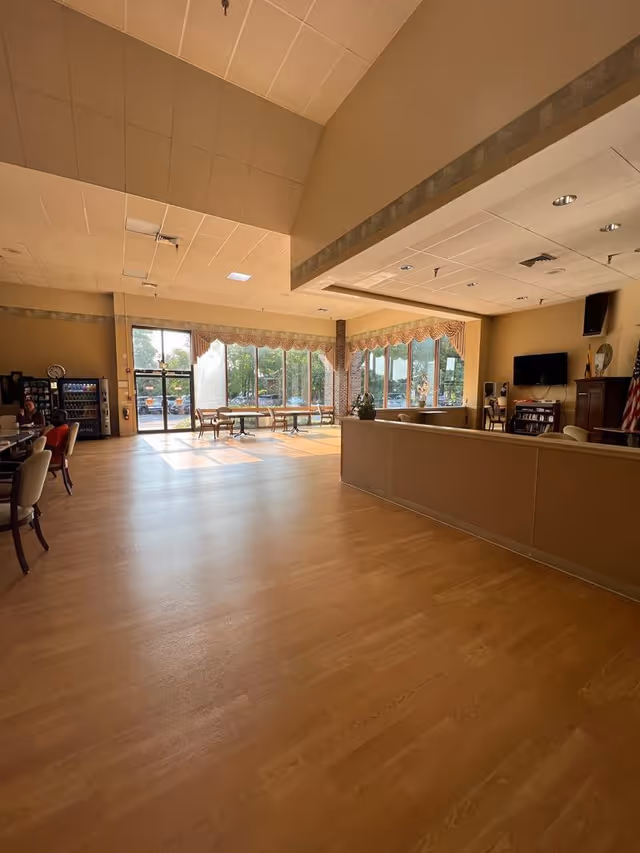 A spacious, well-lit common area with large windows letting in natural light. The room features wooden flooring, several tables and chairs near the windows, a vending machine, a TV mounted on the wall, and an American flag in the corner. The ceiling is high with recessed lighting.