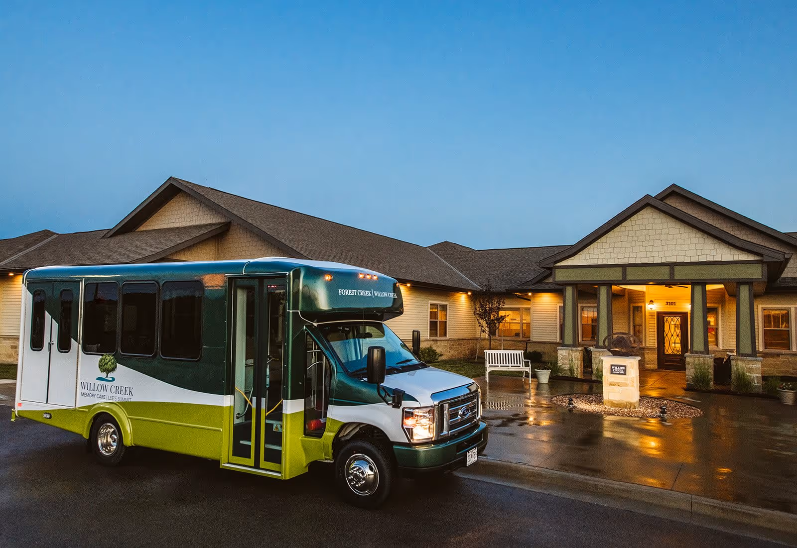 A green and white shuttle bus parked in front of a single-story senior living facility building with a covered entrance and warm exterior lighting during dusk.