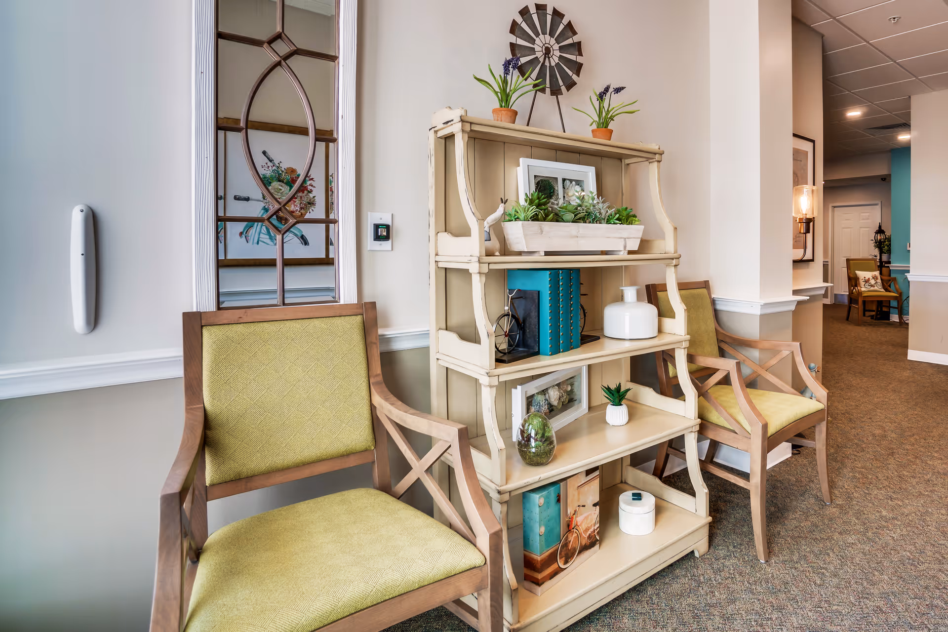 A decorated seating area in a senior living facility hallway with two green upholstered chairs flanking a cream shelving unit filled with plants and decor.