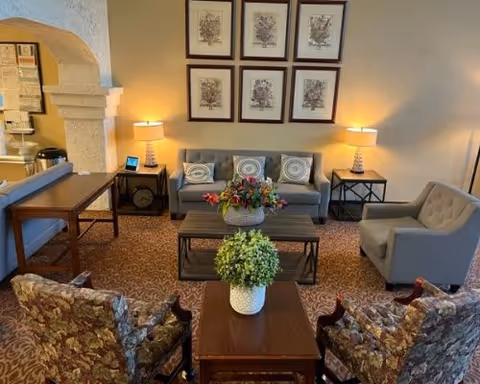 A cozy living room with a gray sofa adorned with three patterned cushions, two floral armchairs, and a gray armchair. There are two side tables with lamps on either side of the sofa, a coffee table with a flower arrangement, and a small table with a green plant in the foreground. The walls are decorated with six framed botanical prints arranged in two rows. The room has warm lighting and a patterned carpet.