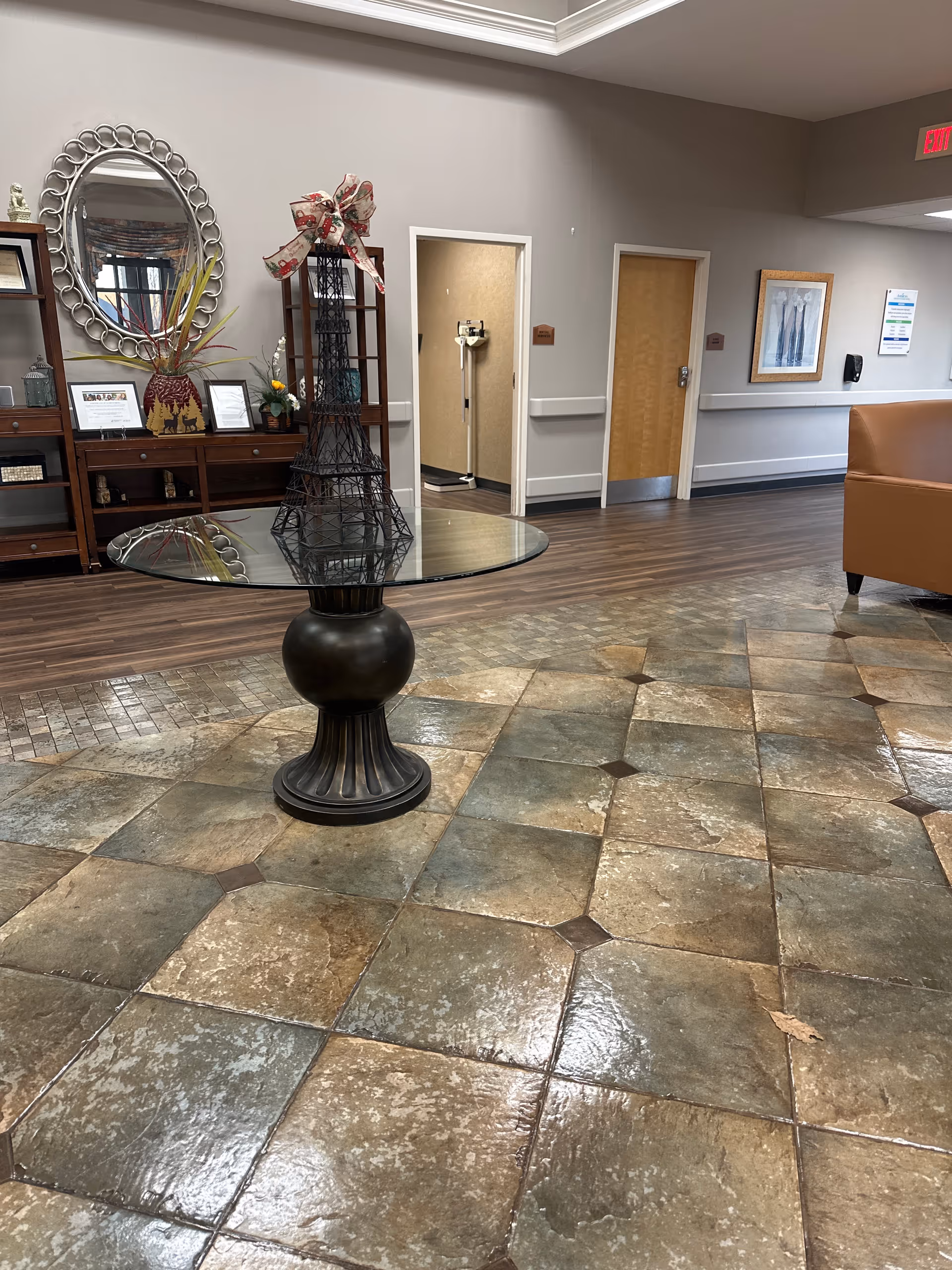 Interior view of a senior living facility lobby with a round glass table featuring a decorative Eiffel Tower sculpture with a bow. The floor has large square tiles in earth tones. In the background, there is a wooden cabinet with a round mirror above it, some framed pictures, and two open doorways leading to other rooms. A brown leather chair is partially visible on the right side.