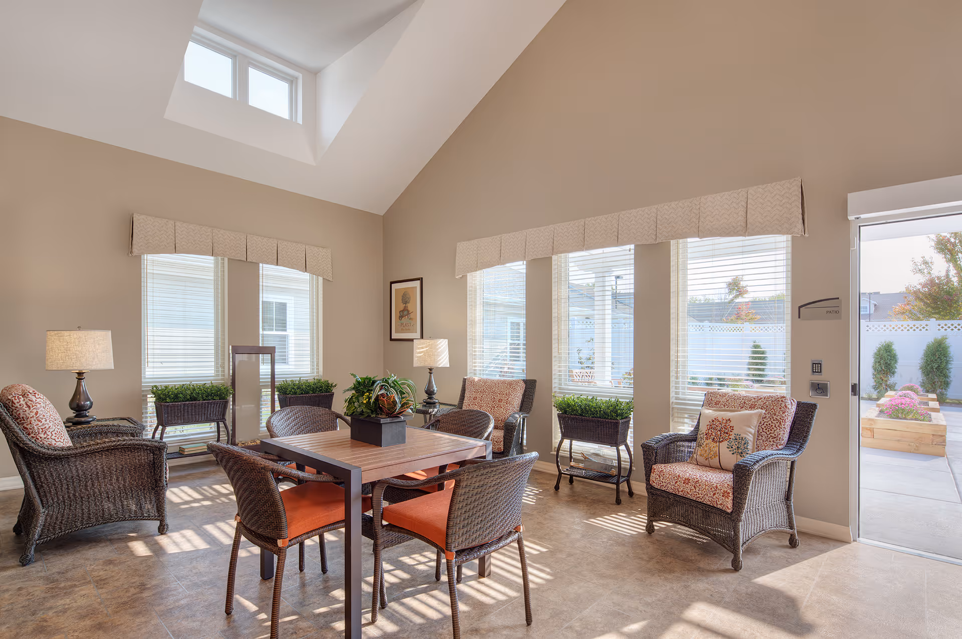 A bright and airy sitting area with wicker chairs featuring patterned cushions and a square wooden table with four wicker chairs with orange cushions. The room has large windows with blinds and valances, letting in natural light. There are two table lamps on side tables, potted plants on stands, and a door leading to an outdoor patio with raised garden beds visible outside.
