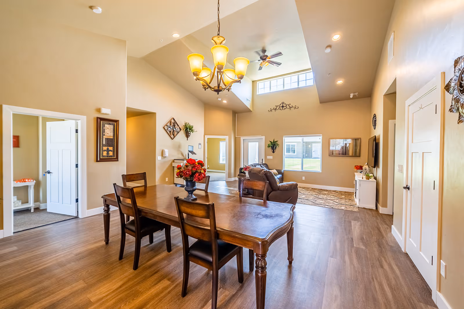 A spacious senior living common area featuring a wooden dining table with four chairs and a vase of red flowers. The room has high ceilings with a chandelier and ceiling fan, beige walls, and large windows letting in natural light. In the background, there are two brown leather sofas, a patterned rug, and wall decorations.