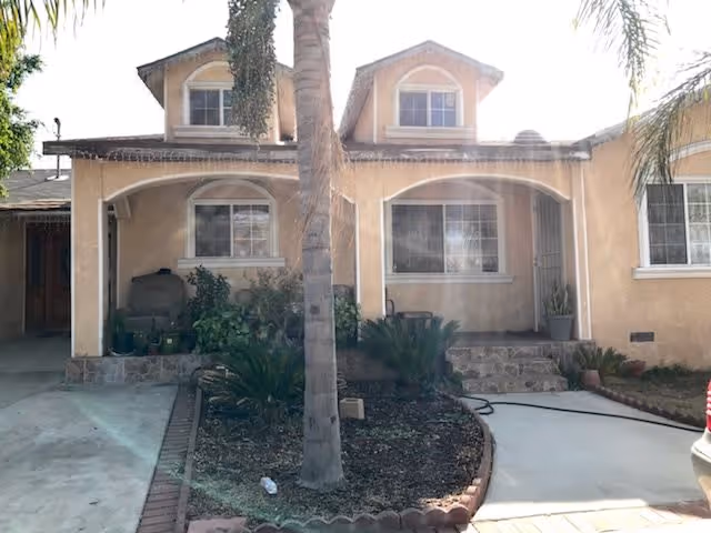 Front exterior view of a beige two-story house with a small garden area featuring a palm tree and various plants. The house has arched windows and a covered porch area with steps leading up to the entrance.