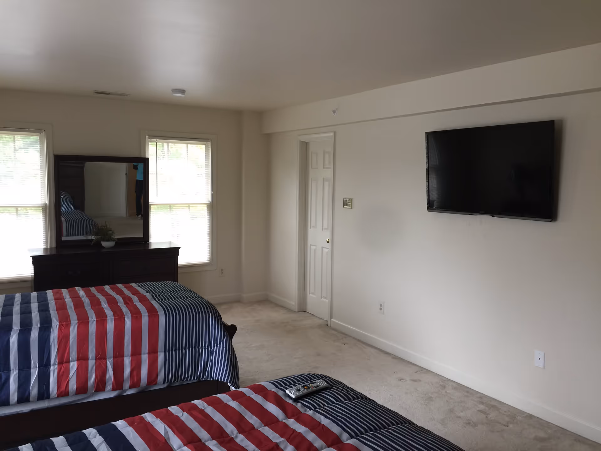 A bedroom with two twin beds covered in red, white, and blue striped bedding. There is a dark wooden dresser with a mirror between two windows with blinds. A flat-screen TV is mounted on the wall opposite the beds. The room has light-colored walls and carpeted floor.