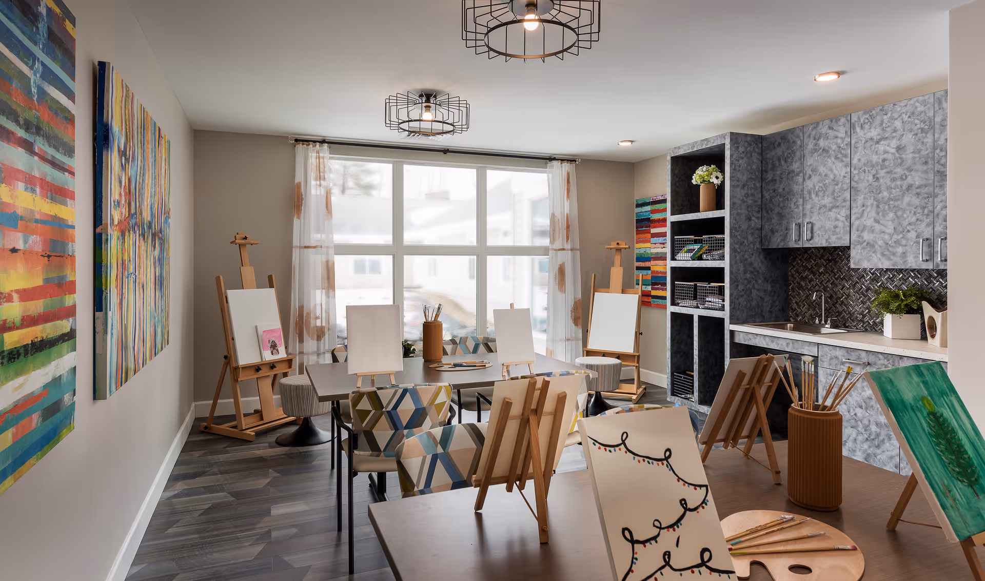 A bright art studio room with several easels holding blank canvases and some with paintings. The room has large windows with sheer curtains, colorful abstract paintings on the walls, patterned chairs around tables, and a kitchenette area with cabinets and a sink.