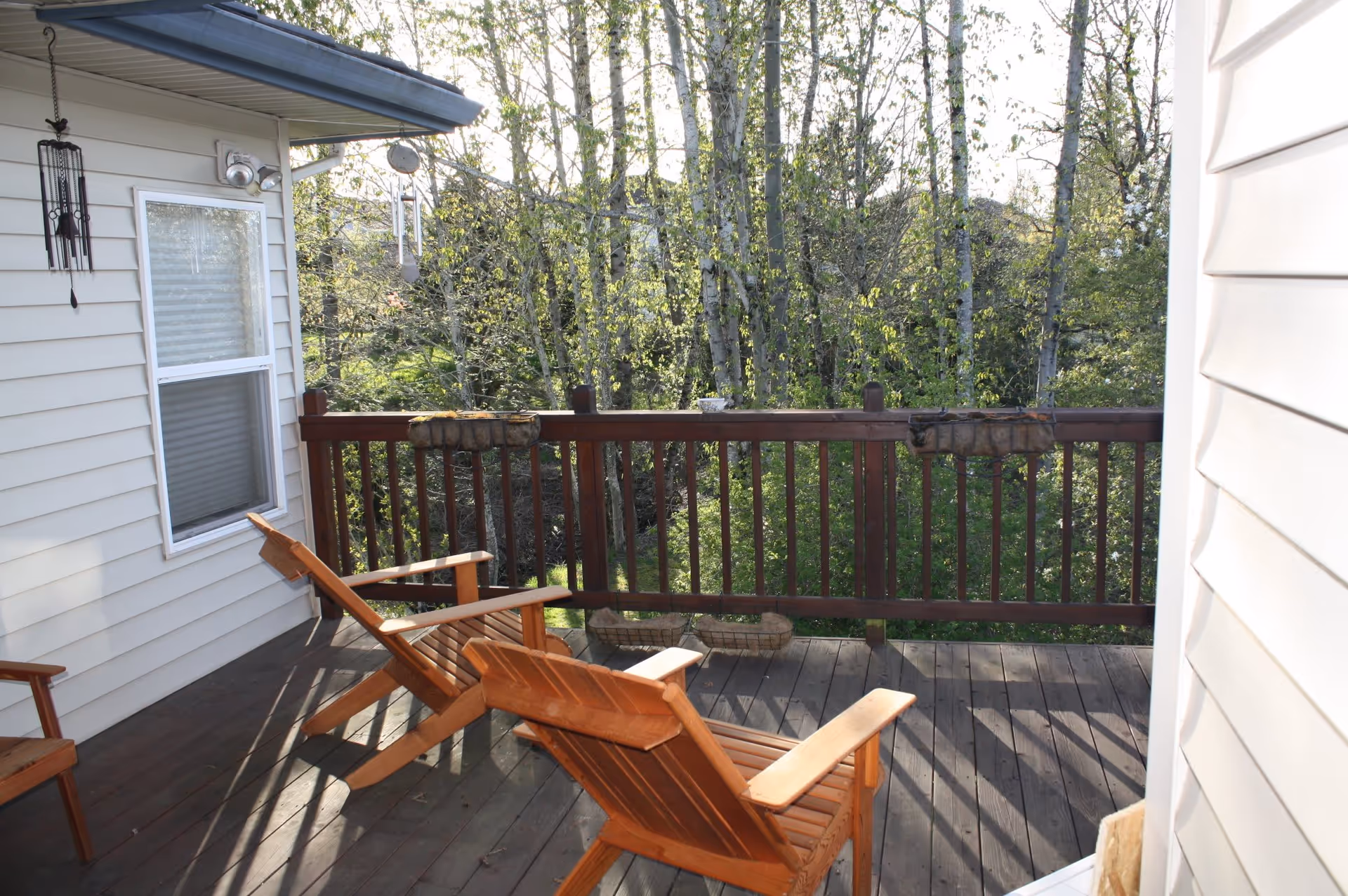Wooden deck with two wooden lounge chairs facing a wooden railing. Behind the railing, there are trees with green leaves. The side of a light-colored house with a window and a wind chime hanging is visible on the left.