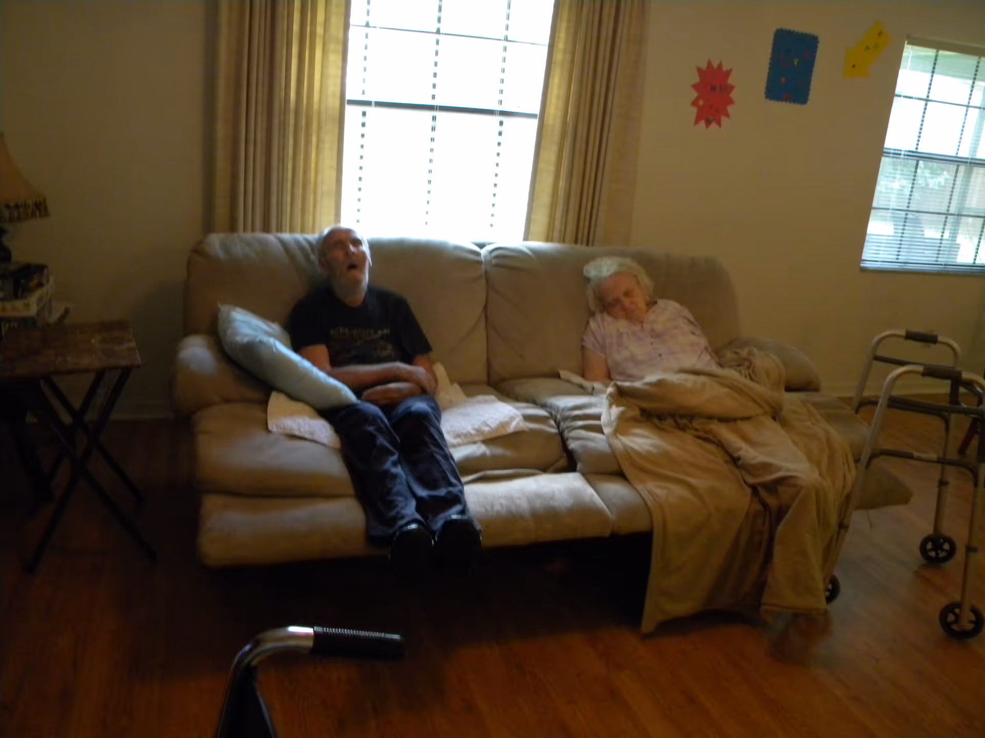 Two elderly individuals sleeping on a beige couch in a room with wooden flooring. There are two windows with blinds and curtains behind the couch. A walker is visible on the right side of the image, and a small table with a lamp and some items is on the left side.