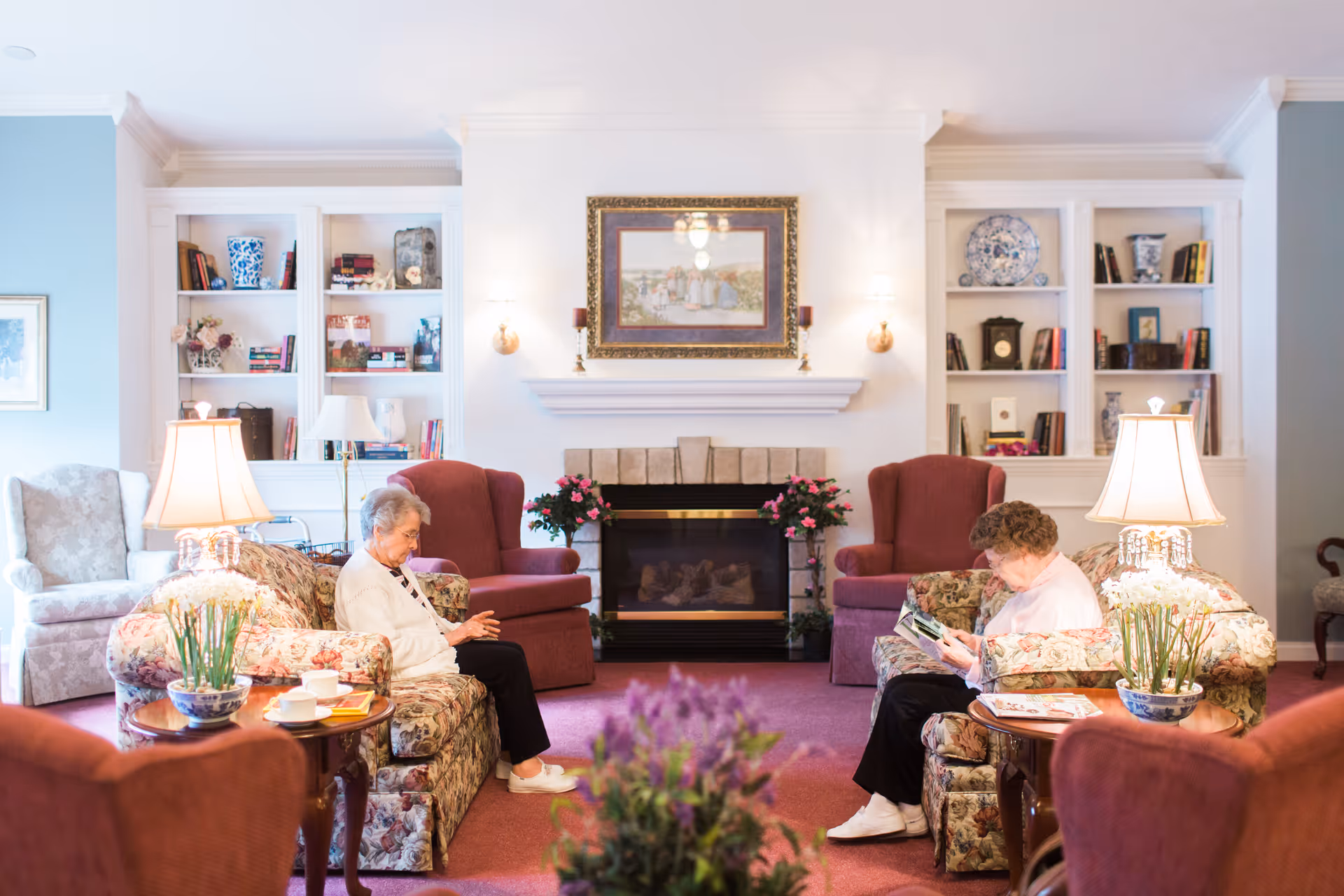 A cozy living room in a senior living facility with two elderly women seated on floral-patterned sofas facing each other. The room features a fireplace with a framed painting above it, built-in white bookshelves filled with books and decorative items, two table lamps on side tables with potted plants, and additional armchairs. The carpet is a warm red color, and the atmosphere is calm and inviting.