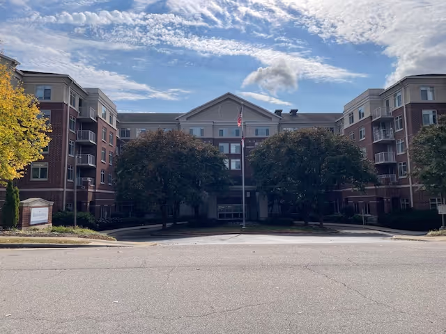 Multi-story brick senior living building facade with a central entrance, an American flag on a flagpole, and trees lining the driveway under a partly cloudy sky.