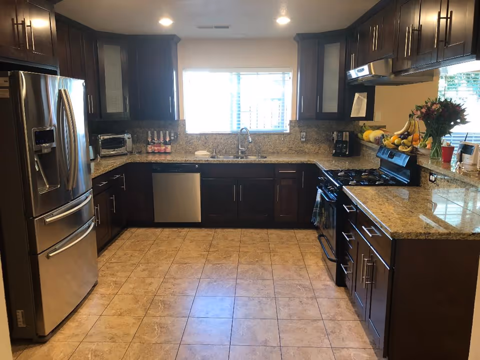 A modern kitchen with dark wood cabinets, granite countertops, and tiled floor. The kitchen features a stainless steel refrigerator, dishwasher, and gas stove with a range hood. There is a window above the sink letting in natural light, and various kitchen appliances and fruit are placed on the countertops.