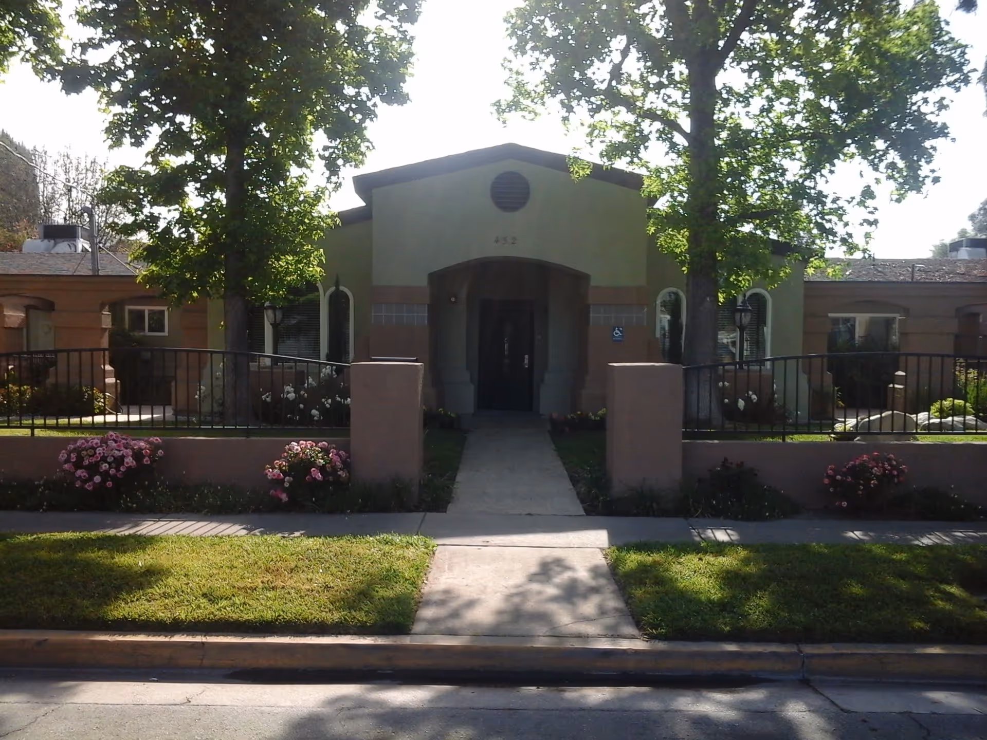 Front exterior view of a single-story building with a central entrance, flanked by two large trees and flower beds along a low wall and sidewalk.