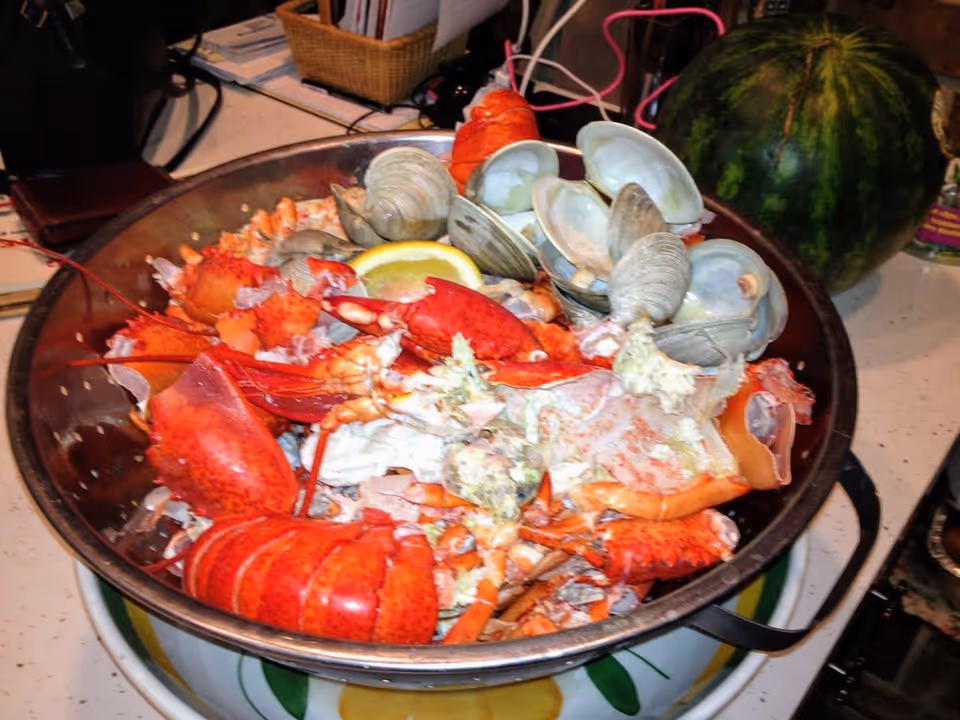 A large metal bowl filled with cooked seafood including lobster claws, crab shells, clams, and a lemon wedge, placed on a kitchen counter with a watermelon and various kitchen items in the background.