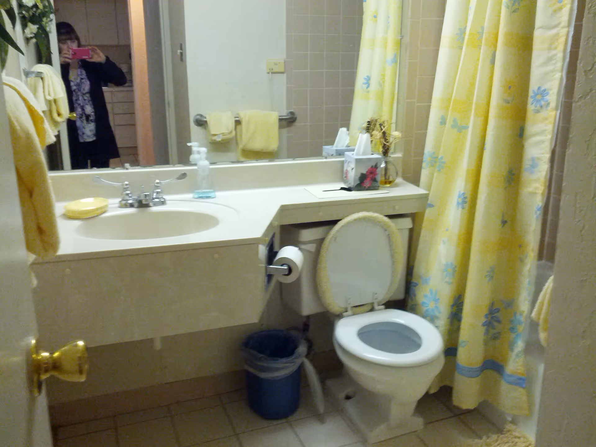 A bathroom with a white sink and countertop, a toilet with a padded seat cover, a yellow and blue floral shower curtain, and yellow towels hanging on the wall. A mirror above the sink reflects a person taking the photo with a pink phone. There is a soap dispenser, a tissue box, and a small vase with dried flowers on the countertop.