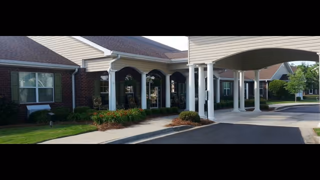 Exterior view of Belvedere Commons of Seneca showing the entrance with white columns, a covered driveway, brick and siding walls, windows, and landscaped greenery including bushes and flowers.