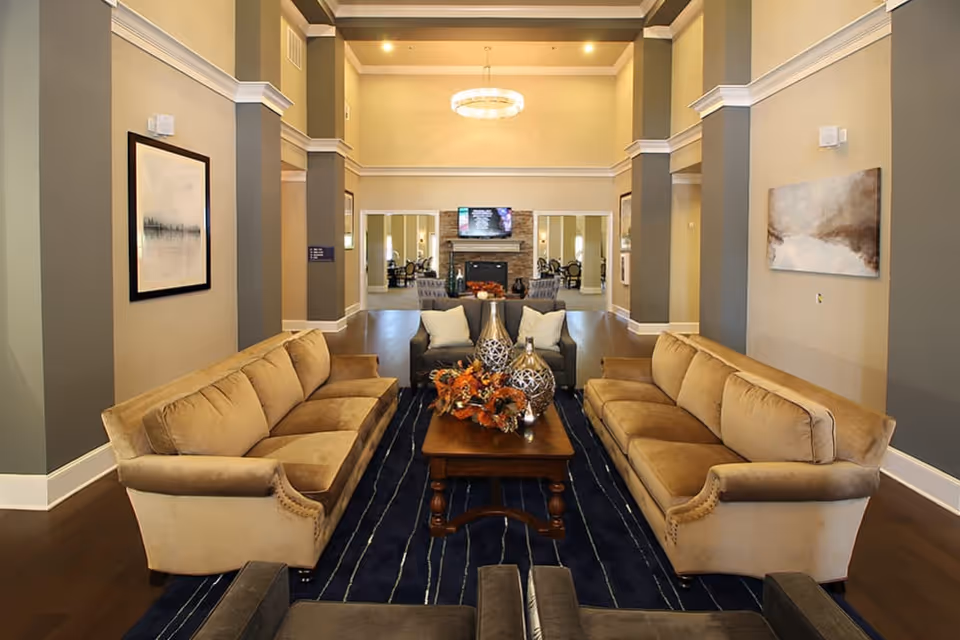 A spacious and elegant living room area with two beige sofas facing each other across a wooden coffee table decorated with vases and autumn-themed floral arrangements. The room features high ceilings with crown molding, a dark blue striped rug, framed artwork on the walls, and a modern chandelier. In the background, there is a fireplace with a mounted TV above it and additional seating visible through an open doorway.