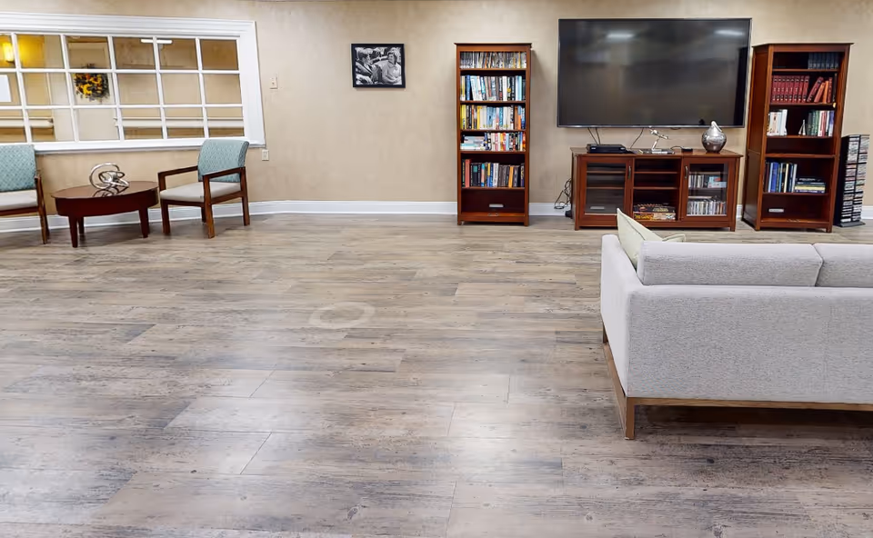 A spacious living room area with wooden flooring, a light gray sofa, two wooden bookshelves filled with books, a large flat-screen TV mounted on the wall above a wooden TV stand, and two armchairs with a small round wooden table between them. There is a window with multiple panes on the left wall and a black and white framed photo hanging on the wall.