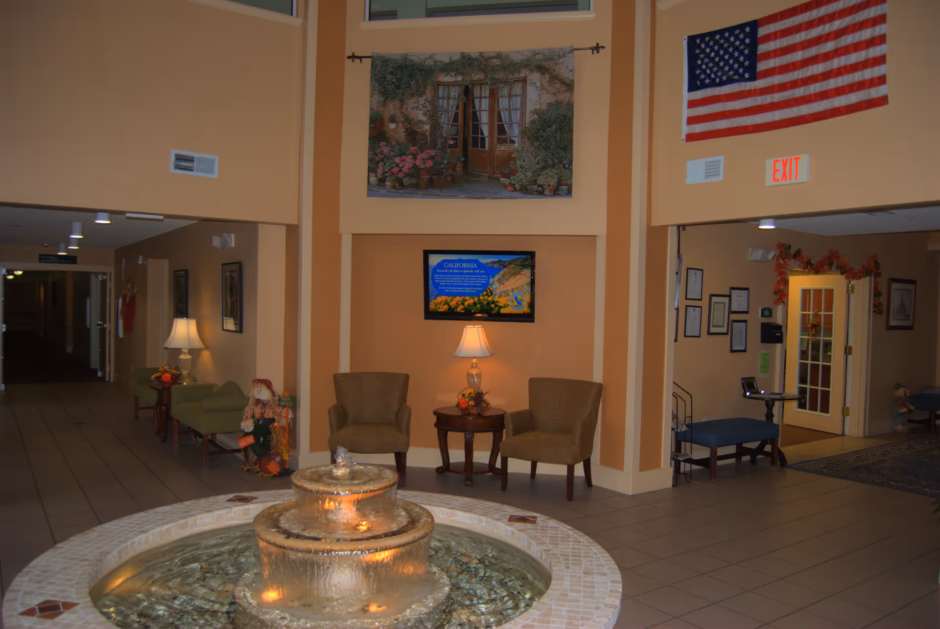 Interior view of a retirement community lobby area with a round water fountain in the foreground. The walls are painted in warm tones and decorated with a large American flag, a framed picture of a garden scene, and a digital display. There are two armchairs and a small table with a lamp between them, as well as additional seating and decorations along the hallway.