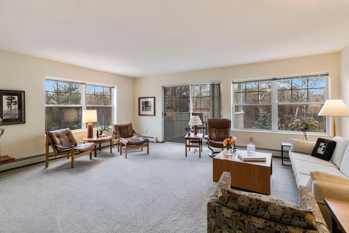 A spacious living room with large windows showing an outdoor view of trees. The room features a beige carpet, a white sofa with a pillow, two brown cushioned chairs, a patterned armchair, a wooden coffee table with flowers and a book, side tables with lamps, and framed artwork on the walls.